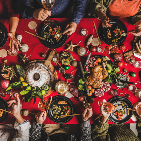 People seated around a red tablecloth enjoying a festive meal with roast chicken, bread, fruit, and various side dishes.