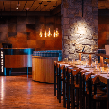 A modern restaurant interior with wooden tables and chairs, stone walls, hanging pendant lights, and a curved booth near an illuminated sign that reads "the source.
