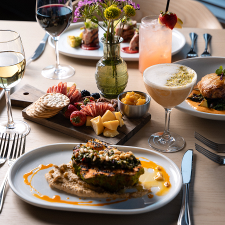 A table set with plated gourmet dishes, assorted cheeses and crackers, fruit, wine, and cocktails, with a vase of flowers as a centerpiece.