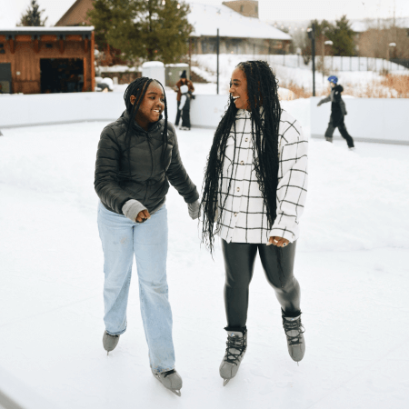 Two people ice skating outdoors, smiling and holding hands, with snow and other skaters in the background.
