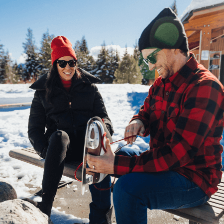 Two people sit on a bench in a snowy outdoor setting; one person is lacing an ice skate while the other looks on and smiles.