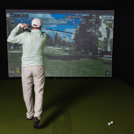 A person in golf attire swings a golf club in front of a large indoor golf simulator screen displaying course information and distances.