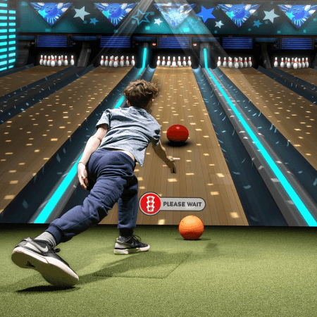 A child bowls a ball down a brightly lit bowling lane, with another ball already rolling and a "please wait" sign visible on the lane.