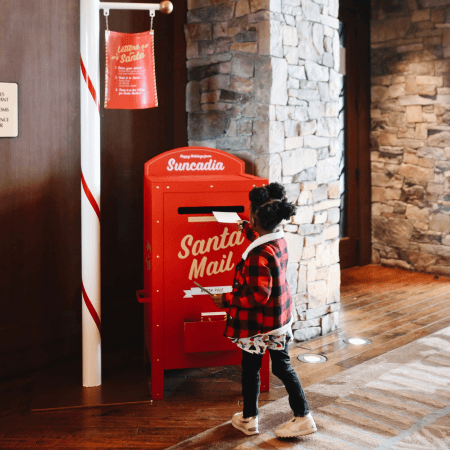 A young child in a plaid jacket stands in front of a red "Santa Mail" mailbox, preparing to send a letter indoors next to a stone wall.