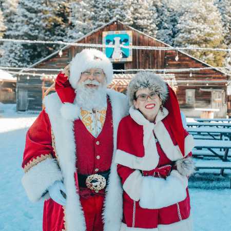 Two people dressed in Santa Claus and Mrs. Claus costumes stand outside in front of a wooden building surrounded by snow.