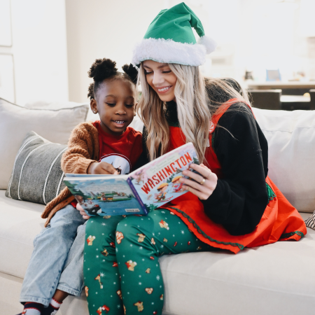 An adult and a child sit on a couch reading a book together. The adult wears a green Santa hat and festive clothes, and the child points at the book and smiles.