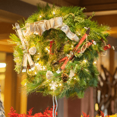 A green Christmas wreath decorated with pinecones, small skis, a gold ribbon, and white lights hangs indoors.