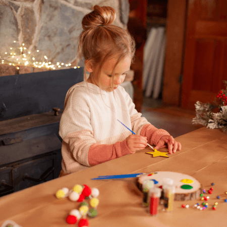 A young girl sits at a table and paints a yellow star-shaped cutout, surrounded by art supplies and craft materials.