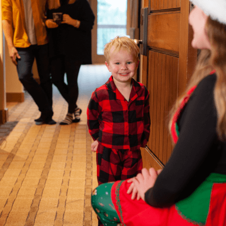 A young child in red plaid pajamas smiles at a woman in festive clothing, while two adults stand in the background of a hallway.