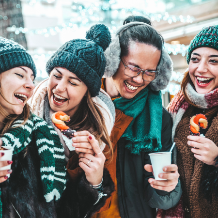 Four people in winter clothing laugh together outdoors, holding hot drinks and snacks, with festive lights in the background.