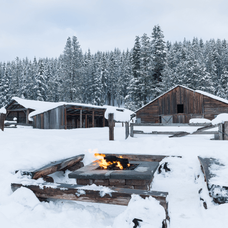 A fire burns in a stone pit surrounded by snow, with several wooden cabins and tall pine trees in the background under a cloudy winter sky.
