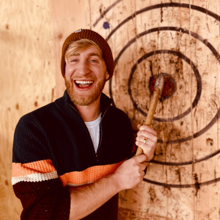 A man smiles while holding an axe against the bullseye of a wooden target at an axe-throwing venue.