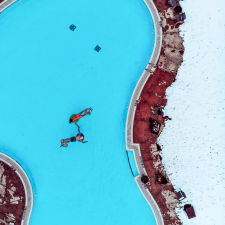 Aerial view of two people in colorful swimsuits floating in a pool next to a snowy, partially cleared pool deck.