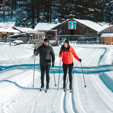 Two people cross-country skiing on groomed snow trails with a wooden cabin and trees in the background.