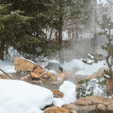Two people sit in an outdoor hot spring surrounded by snow-covered rocks and trees, with steam rising and snow falling gently around them.