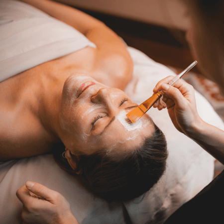 A person lying on a spa bed receives a facial treatment as a brush is used to apply a white mask to their forehead.