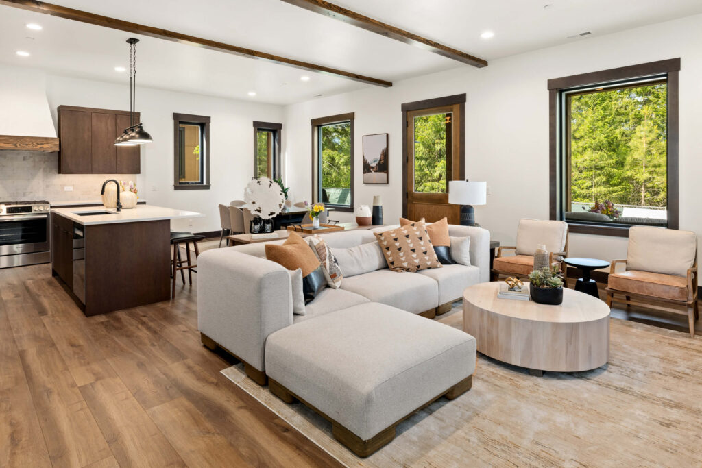 Modern open-concept living room and kitchen with wood floors, neutral furniture, dark cabinets, large windows, and natural light.