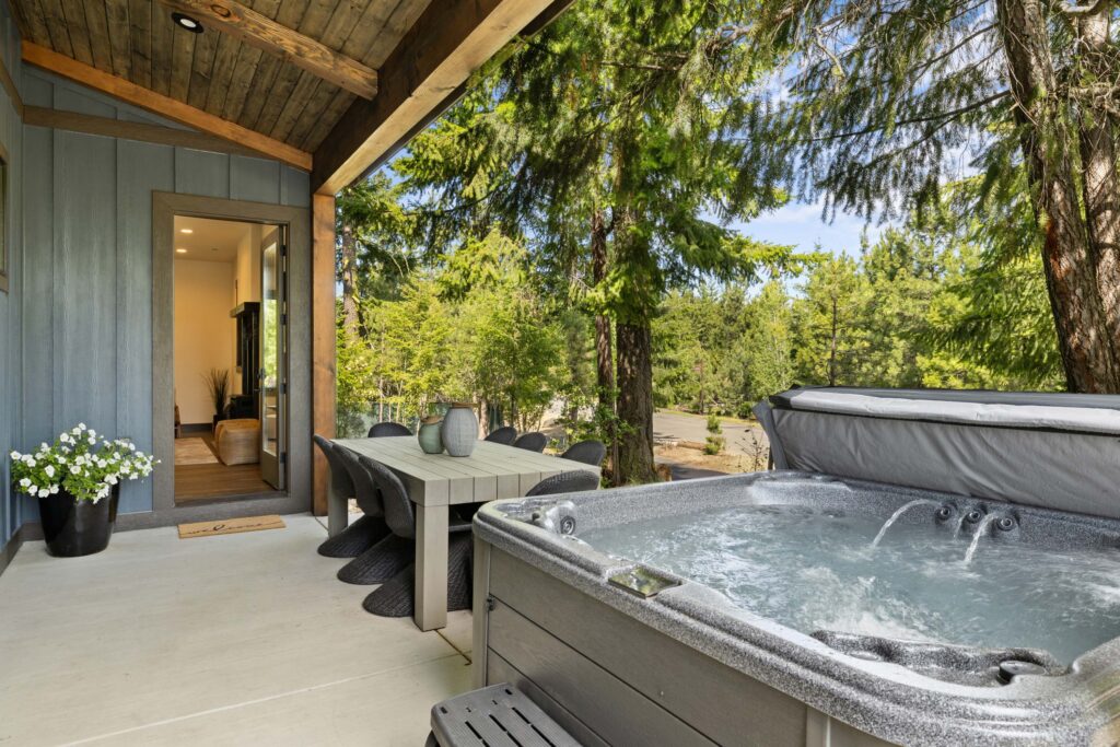 A covered patio with a table and chairs, potted flowers, and a hot tub, overlooking a wooded area with tall green trees.