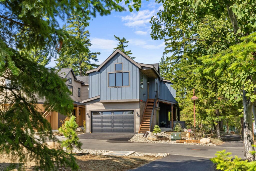 Two-story modern house with blue-gray siding, a double garage, and wooden stairs, surrounded by trees and landscaped yard on a sunny day.
