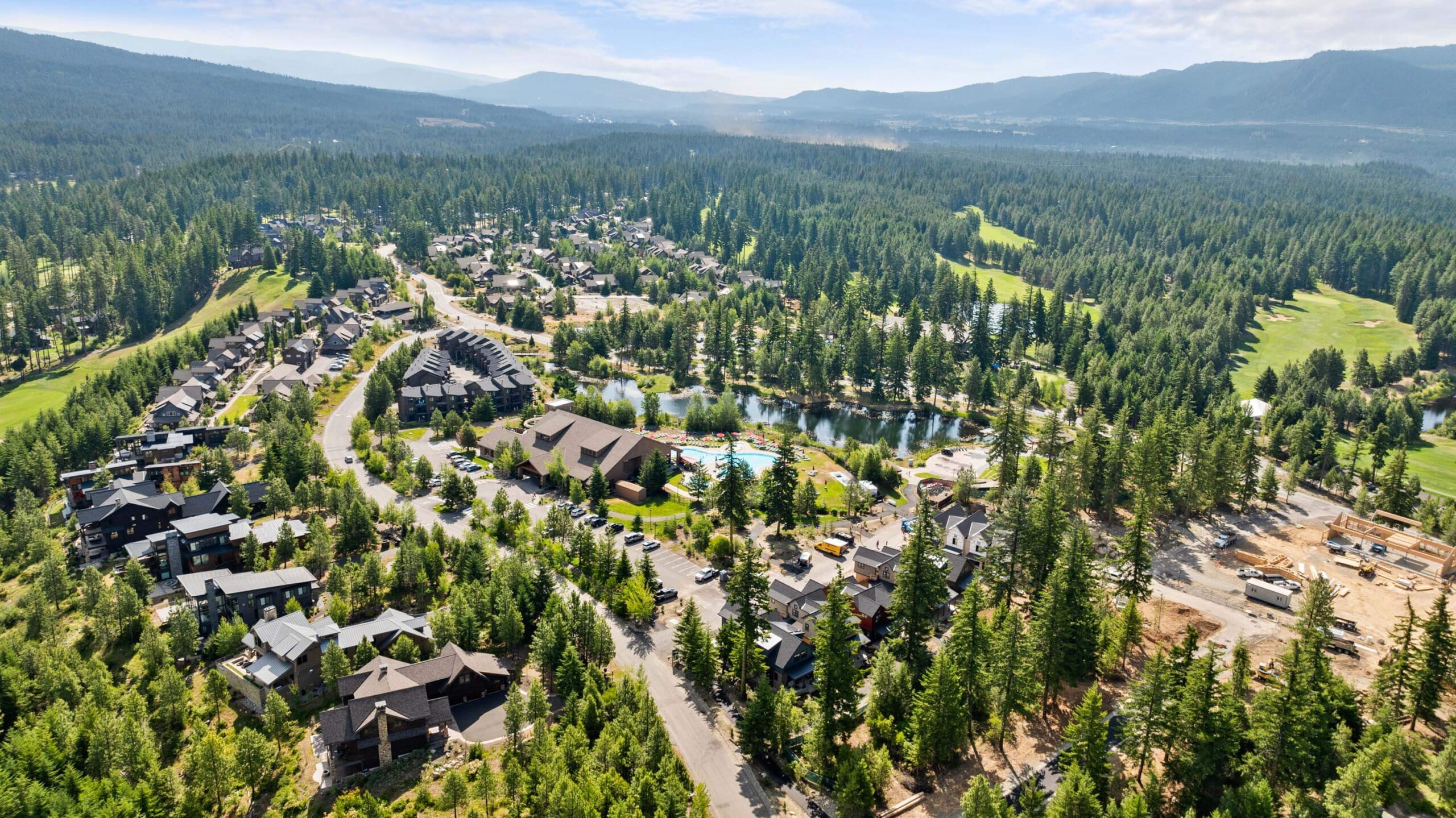Aerial view of a residential neighborhood surrounded by dense forest, with houses, roads, a small lake, and mountains in the background.