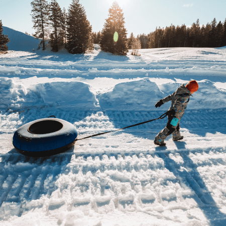 A child wearing a snowsuit and helmet pulls an inner tube across a snowy landscape with trees and sunlight in the background.