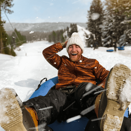 A person in a brown jacket and white knit hat is tubing down a snowy hill, smiling with legs outstretched, surrounded by snow and trees.