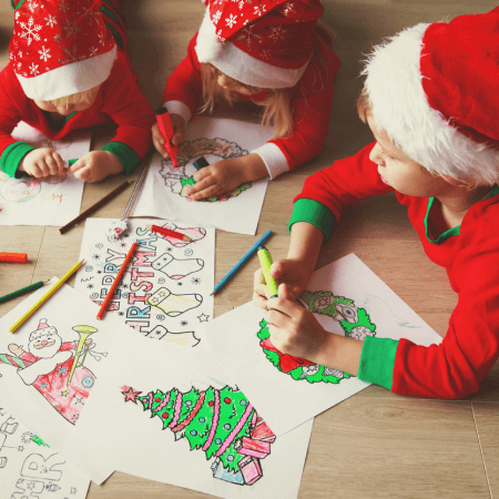 Three children in Santa hats color Christmas-themed drawings with markers and colored pencils on a wooden floor.