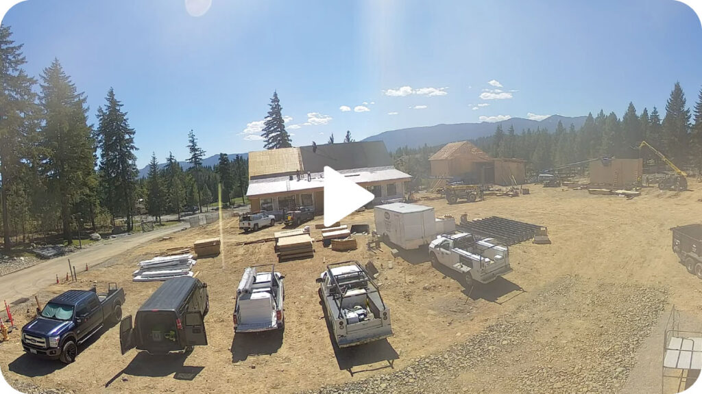 A construction site with several vehicles, building materials, and partially built structures surrounded by trees under a clear sky.