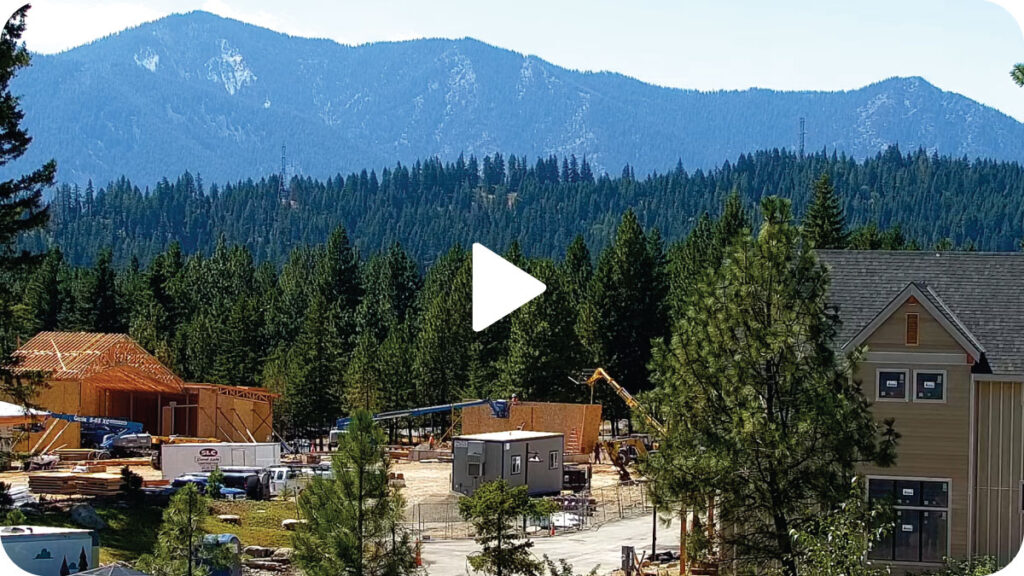 Construction site with several wooden structures, construction vehicles, and equipment surrounded by pine trees, with mountains visible in the background.