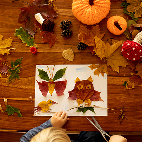 A child making art with autumn leaves and paper on a wooden table, surrounded by leaves, pinecones, yarn pumpkins, and a stuffed mushroom.