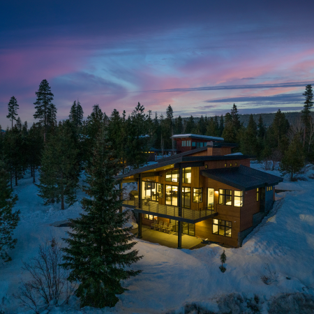 Modern house with large windows lit up at dusk, surrounded by snow and pine trees under a purple and blue sky.