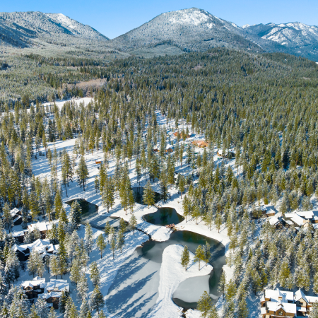Aerial view of snow-covered houses, frozen ponds, and pine trees in a mountain valley on a clear winter day.