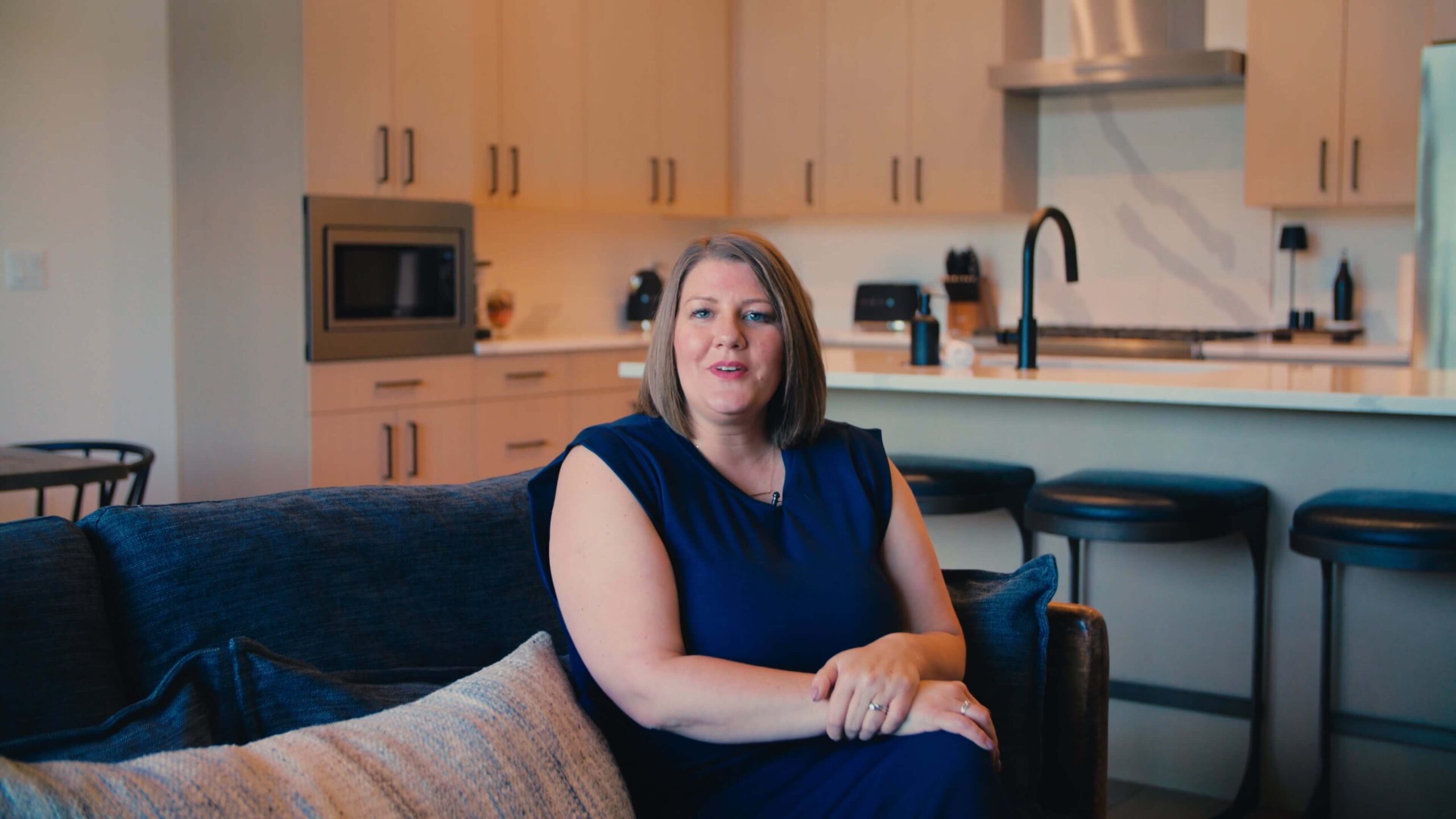 A woman in a blue dress sits on a dark sofa in a modern kitchen with light cabinets, a microwave, and bar stools visible in the background.