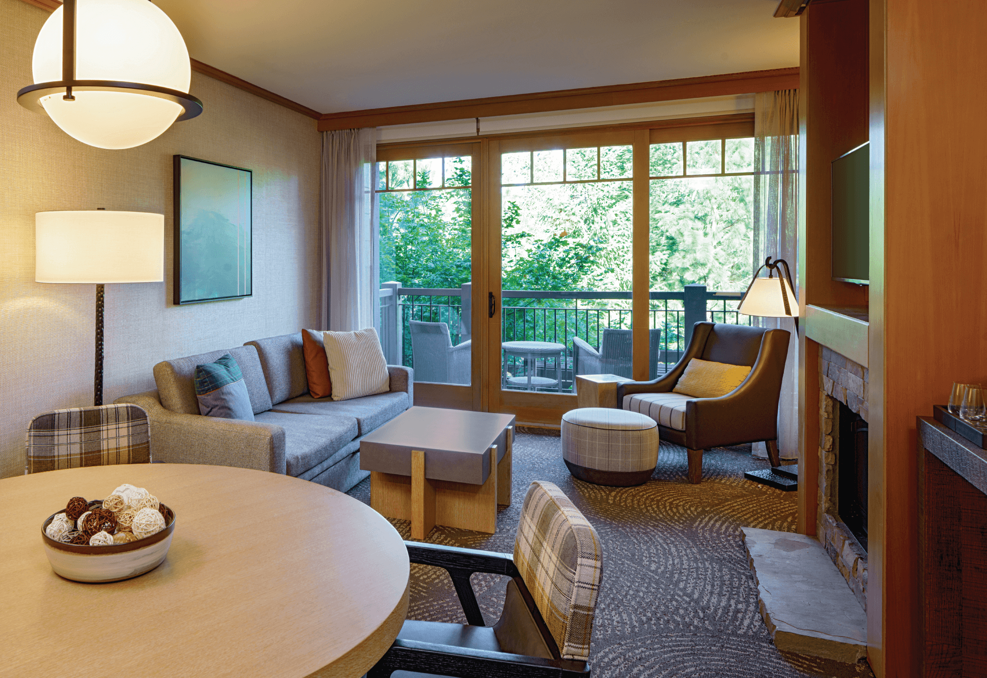 Modern hotel suite living area with a sofa, armchair, round dining table, fireplace, wall-mounted TV, and glass doors leading to a balcony overlooking greenery.