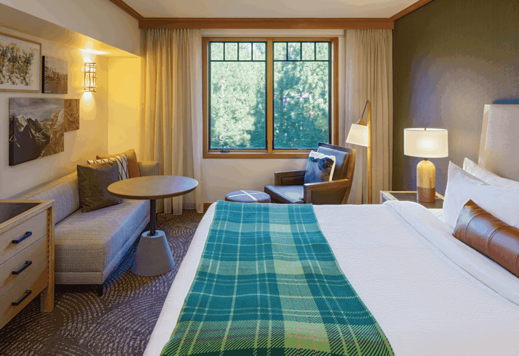 A hotel room with a bed, green plaid blanket, sofa, round table, armchair, lamps, and a large window with closed curtains showing green trees outside.