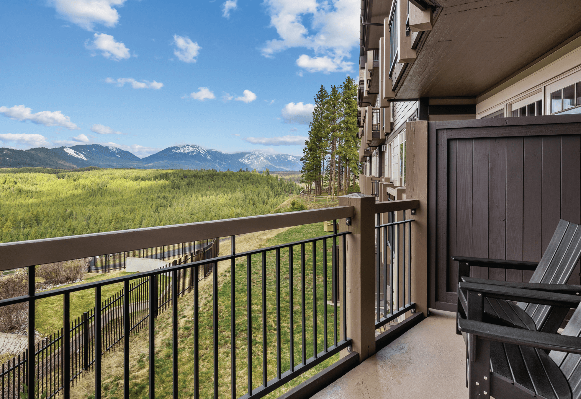 A balcony with two black rocking chairs overlooks a green valley, forest, and distant mountains under a blue sky with scattered clouds.