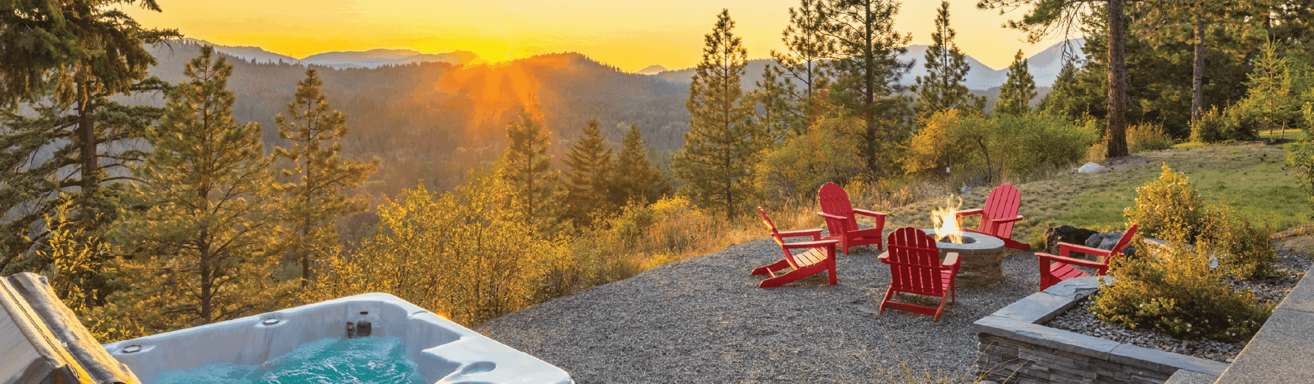 A hot tub and red Adirondack chairs surround a fire pit overlooking a scenic mountain landscape at sunset.
