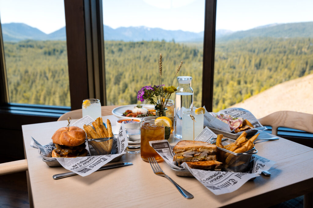 A table set with burgers, fries, sandwiches, drinks, and plates of food, positioned by large windows overlooking a forested mountain landscape.
