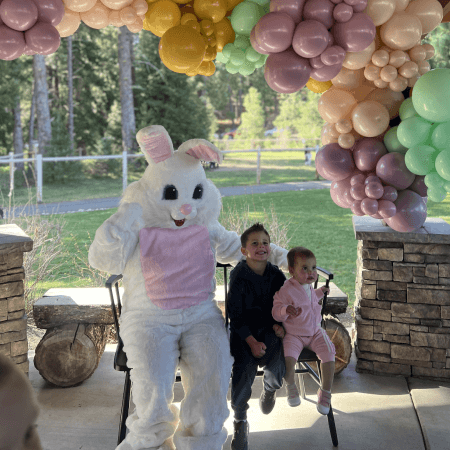 Two children sit on chairs next to a person in a white Easter Bunny costume under a pastel balloon arch outdoors, with trees and grass visible in the background.