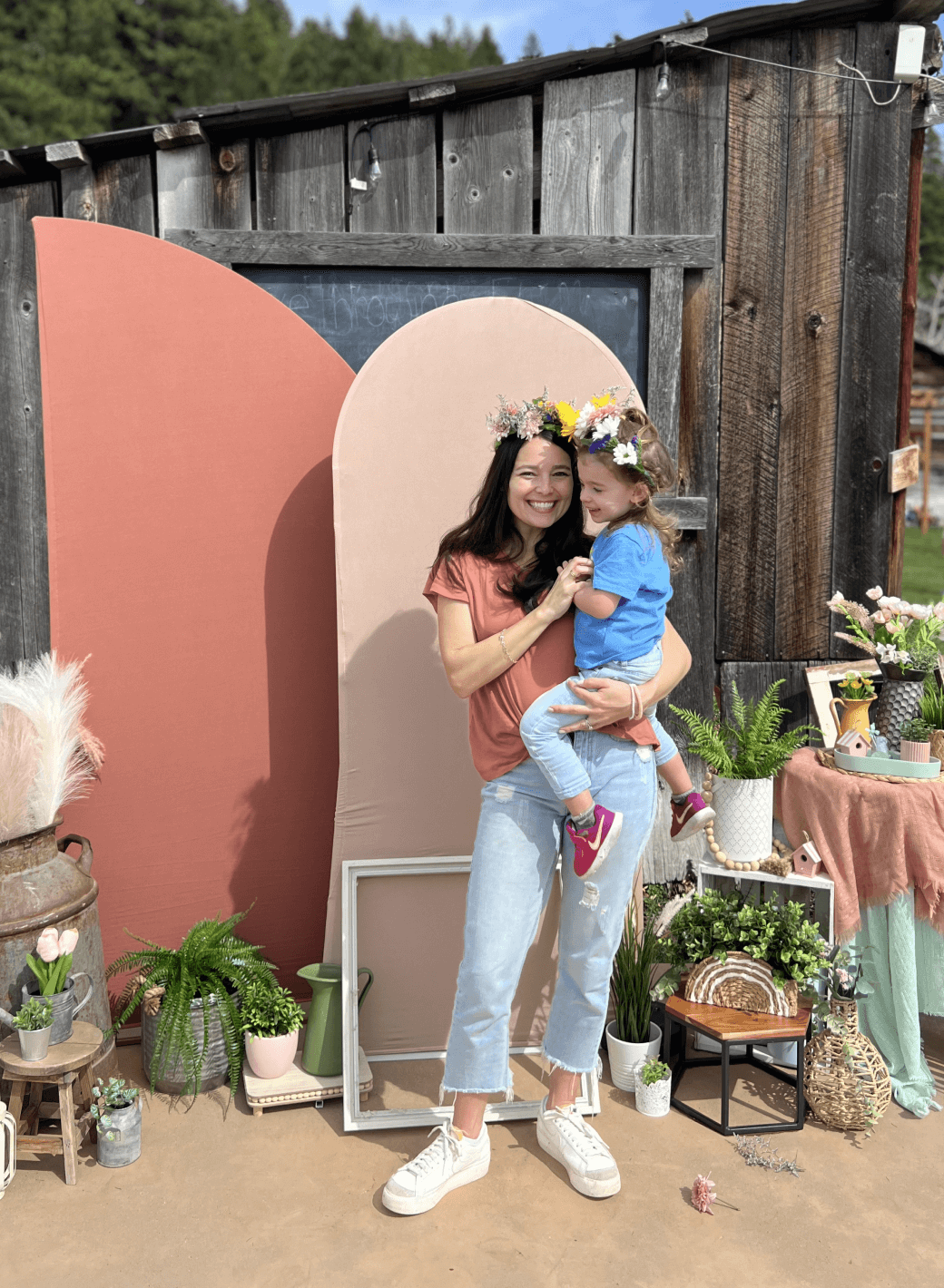 A smiling woman wearing a flower crown holds a young child in front of a rustic wooden backdrop decorated with plants, flowers, and pastel props outdoors.