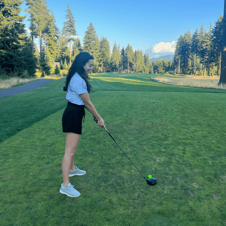 A woman stands on a golf course, preparing to tee off. She holds a golf club and looks toward the fairway, with tall trees and blue sky in the background.