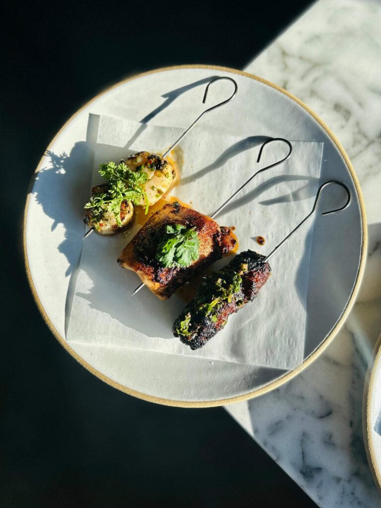 A white plate from Eldr at Suncadia Social holds three pieces of grilled meat and vegetables on skewers, each garnished with fresh herbs, placed on a square sheet of parchment paper on a marble table in natural sunlight.