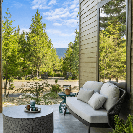 A cozy outdoor patio with a cushioned sofa and round stone table overlooks a scenic landscape of pine trees and distant mountains under a partly cloudy blue sky.