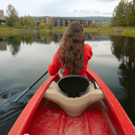 A person with long brown hair wearing a red jacket paddles a red kayak on a calm lake, facing away from the camera. Trees and buildings are visible in the background.