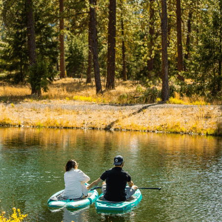 Two people sit on inflatable paddle boards in a calm, forested lake, facing away. Sunlight filters through tall pine trees, illuminating the shore and reflecting off the water’s surface.