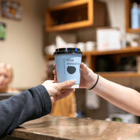 A person hands a takeaway coffee cup labeled the source to another person over a counter in a café, with shelves and blurred café items in the background.