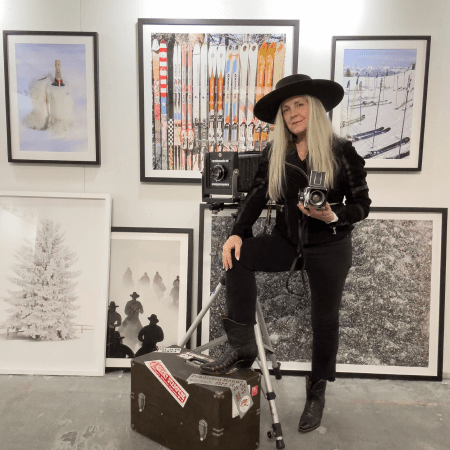 A woman in black clothing and a wide-brimmed hat holds a vintage camera, posing with one foot on a tripod. She is surrounded by framed photos of wintry scenes and a suitcase covered in stickers.