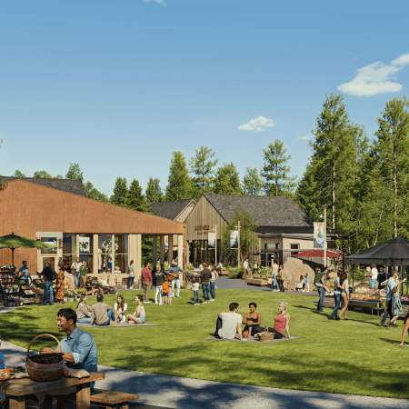 People gather on a sunny day in a green open space with modern buildings, picnic tables, market stalls, and outdoor seating, surrounded by trees under a blue sky. Some are eating, talking, or relaxing on the grass.