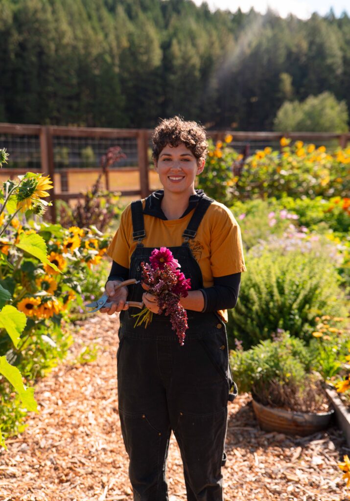 A person in black overalls and a yellow shirt smiles while holding a bouquet of colorful flowers and gardening shears in a vibrant garden—capturing the joyful spirit of Spring events at Suncadia, with sunflowers, green plants, and trees in the background.