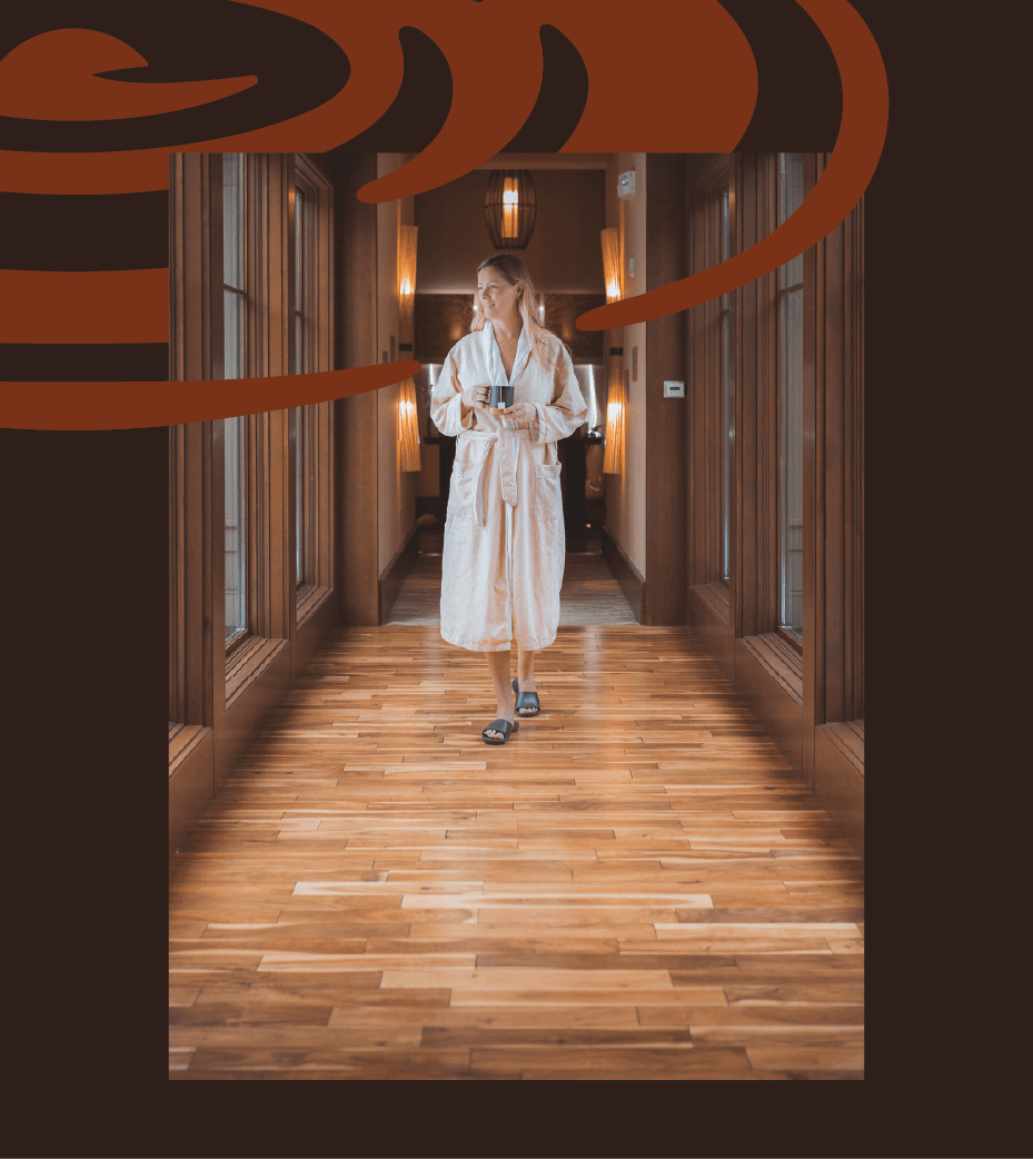 A woman in a white robe walks down a wooden hallway holding a cup, with warm lighting and large windows. Artistic brown swirl graphics overlay the top of the image.